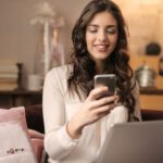 Mobile App - Woman Sitting on Sofa While Looking at Phone With Laptop on Lap