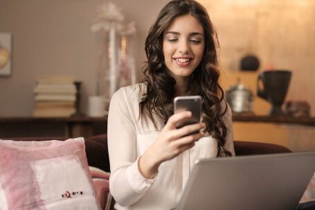 Mobile App - Woman Sitting on Sofa While Looking at Phone With Laptop on Lap