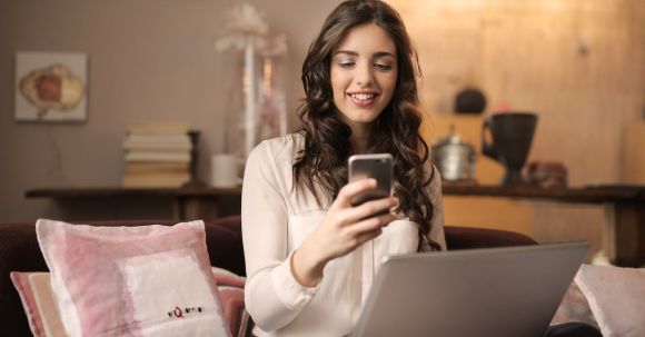 Mobile App - Woman Sitting on Sofa While Looking at Phone With Laptop on Lap